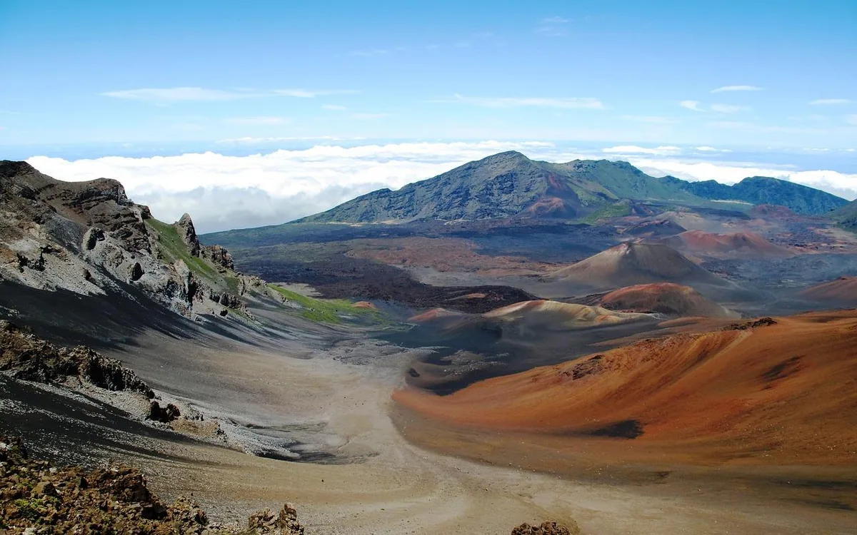 A pale sandy crater floor with cinder cones standing on it and a green ridge behind