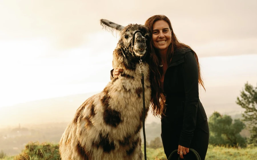 A smiling woman standing beside a llama on a grassy slope