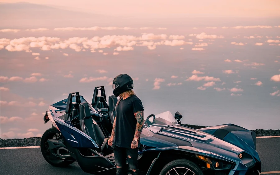 A driver in a helmet standing beside a silver three-wheeler at the roadside